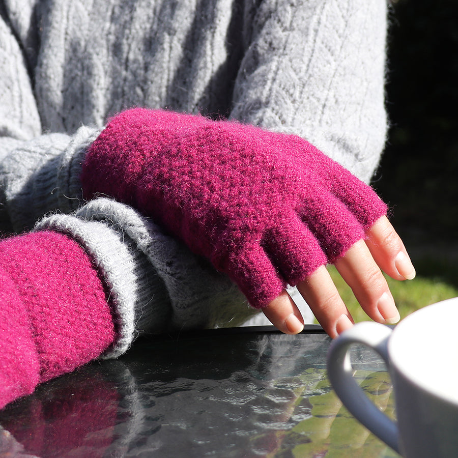 A person wearing waffle-textured magenta fingerless gloves and a grey jumper rests their hands on a glass table beside a white cup, with blurred greenery in the background.