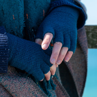 A person wearing blue vegan fingerless gloves and a textured coat stands outdoors, hands clasped together.