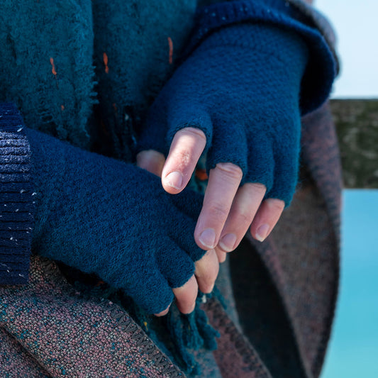 A person wearing blue vegan fingerless gloves and a textured coat stands outdoors, hands clasped together.