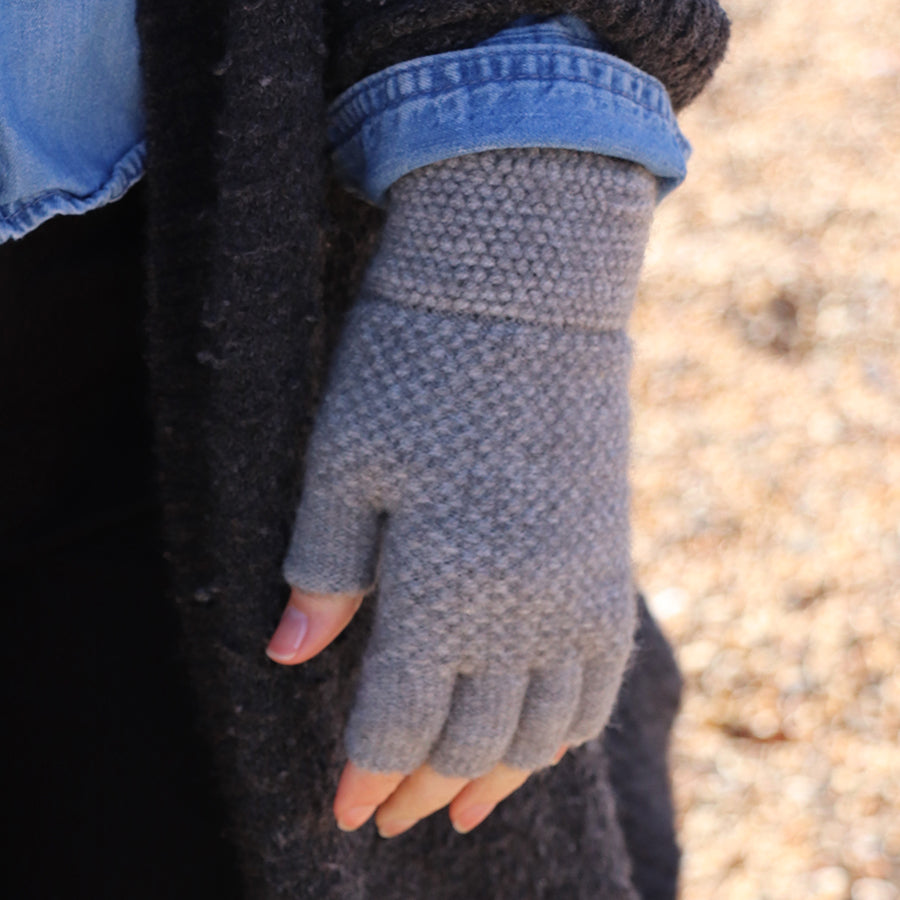 A person stands outdoors in a dark coat, their hand resting at their side, displaying a grey, textured fingerless glove—an ideal choice for autumn accessories.