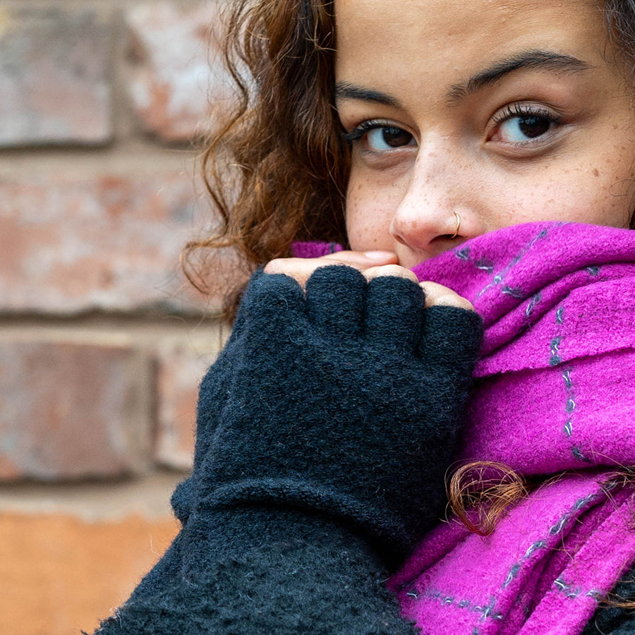 A person with curly hair, sporting a nose ring, black vegan fingerless gloves, and a vibrant magenta scarf stands in front of a brick wall.