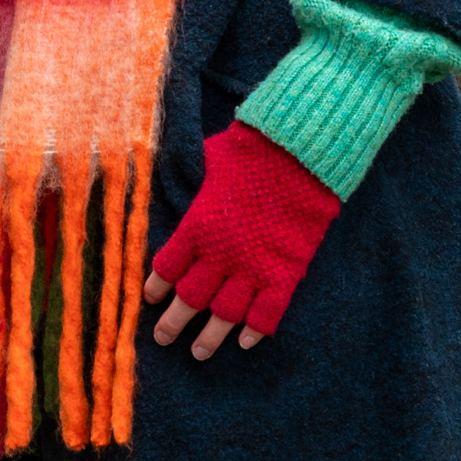 A person wearing red vegan fingerless gloves, a bright green jumper, and a multicoloured fringed scarf over a dark coat—ideal for showcasing striking autumn winter accessories.