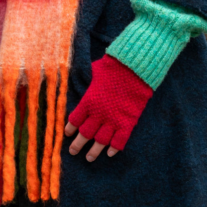 A person wearing red vegan fingerless gloves, a bright green jumper, and a multicoloured fringed scarf over a dark coat—ideal for showcasing striking autumn winter accessories.