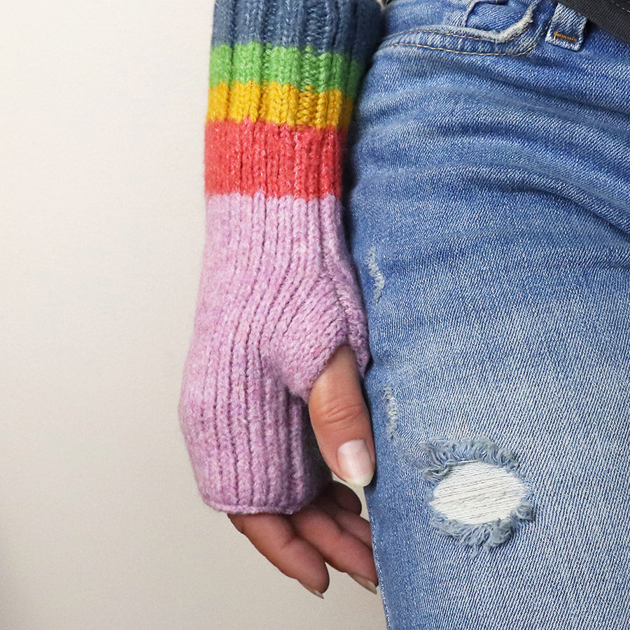 A person wearing distressed blue jeans and knitted, cruelty-free mauve rainbow mittens with fingerless cuffs.