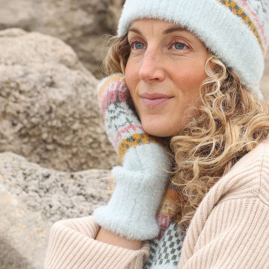 A person with curly blonde hair wears cosy accessories—a knitted hat, scarf, and mittens in pastel colours—touching their face and standing in front of rocks.