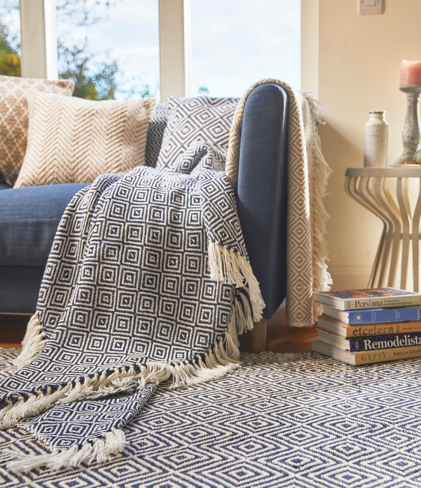 A blue sofa with patterned cushions, a sustainable throw, and a matching rug; a stack of magazines and a candle sit on a side table in this well-lit sitting room.