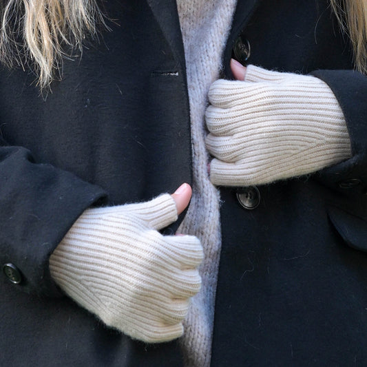 Close-up of a person in a black coat, light grey jumper, and cosy ribbed beige fingerless gloves, with their hands holding the coat lapels.