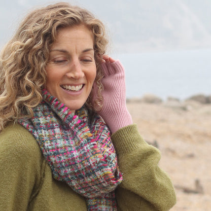 A woman with curly hair smiles while standing outdoors, dressed in a green jumper, multicoloured scarf, and dusky pink fingerless gloves made from recycled materials, near a body of water.