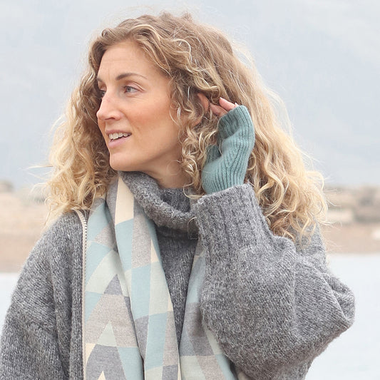 A woman with curly blonde hair in a grey jumper, patterned scarf, and green vegan fingerless gloves stands outside, embracing eco-friendly fashion as she gazes to the side.