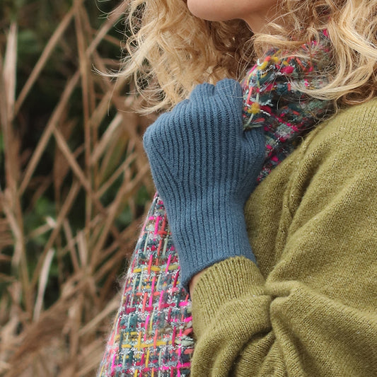 A person in a green jumper, multicoloured scarf, and blue fingerless gloves made from vegan recycled material stands outdoors with dried plants in the background.
