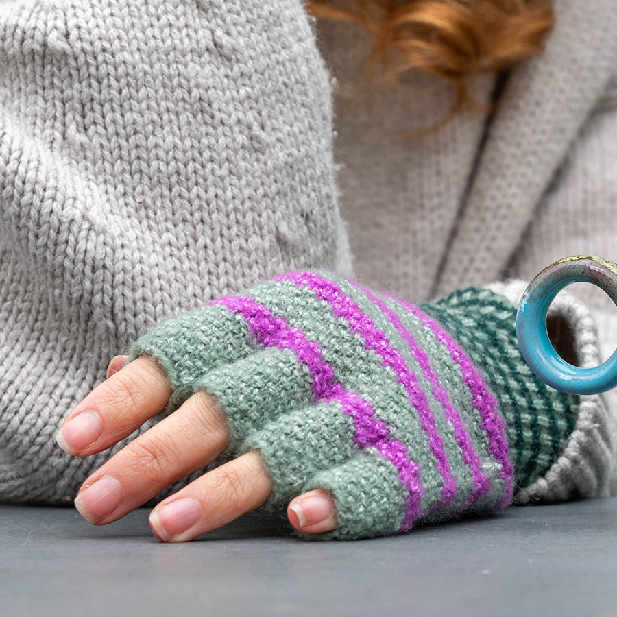 A person wearing a grey jumper and fingerless gloves made from a recycled blend with green and purple stripes rests their hand on a surface.