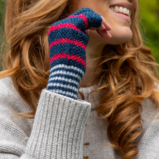 A person with wavy auburn hair wears a grey knitted jumper and vegan fingerless gloves with navy blue and red stripes, smiling outdoors.