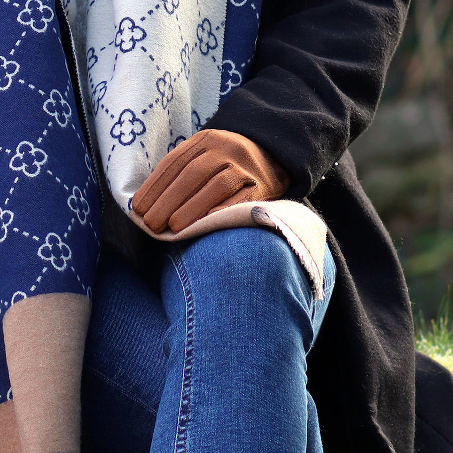 A person wearing vegan-friendly gloves rests their hand on their knee, dressed in blue jeans, a patterned scarf, and a black coat—ideal winter fashion accessories for the season.