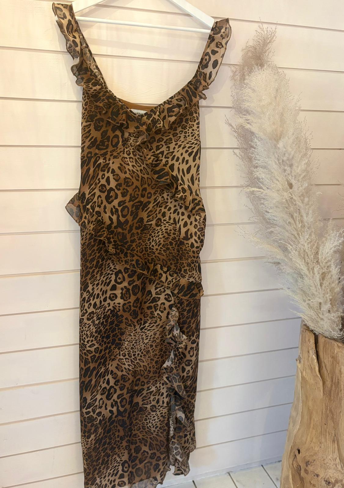 A sleeveless, ruffled leopard print dress in size 12 hangs on a white hanger against a light wood panel wall. Pampas grass in a wooden vase can be seen to the right.