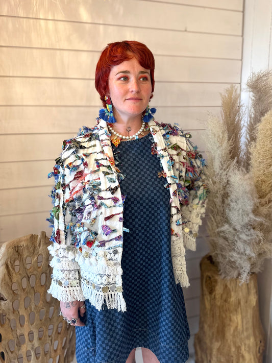 A person with short red hair is wearing a blue patterned dress and a white textured hippy jacket adorned with colourful fabric strips, standing indoors beside beige pampas grass and wooden decor.