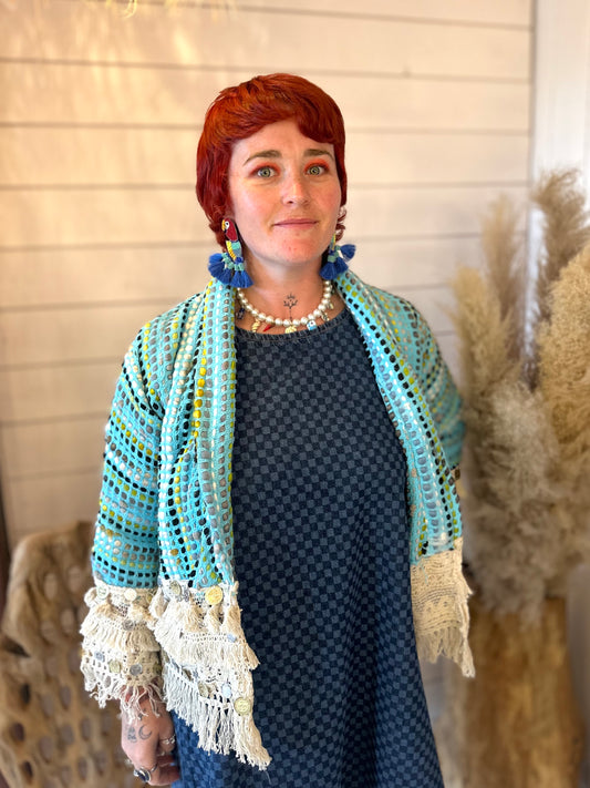 A person with short red hair is wearing a blue chequered dress, a light blue fringed shawl, statement earrings, and a beaded necklace, standing indoors near pampas grass and TBC décor elements.