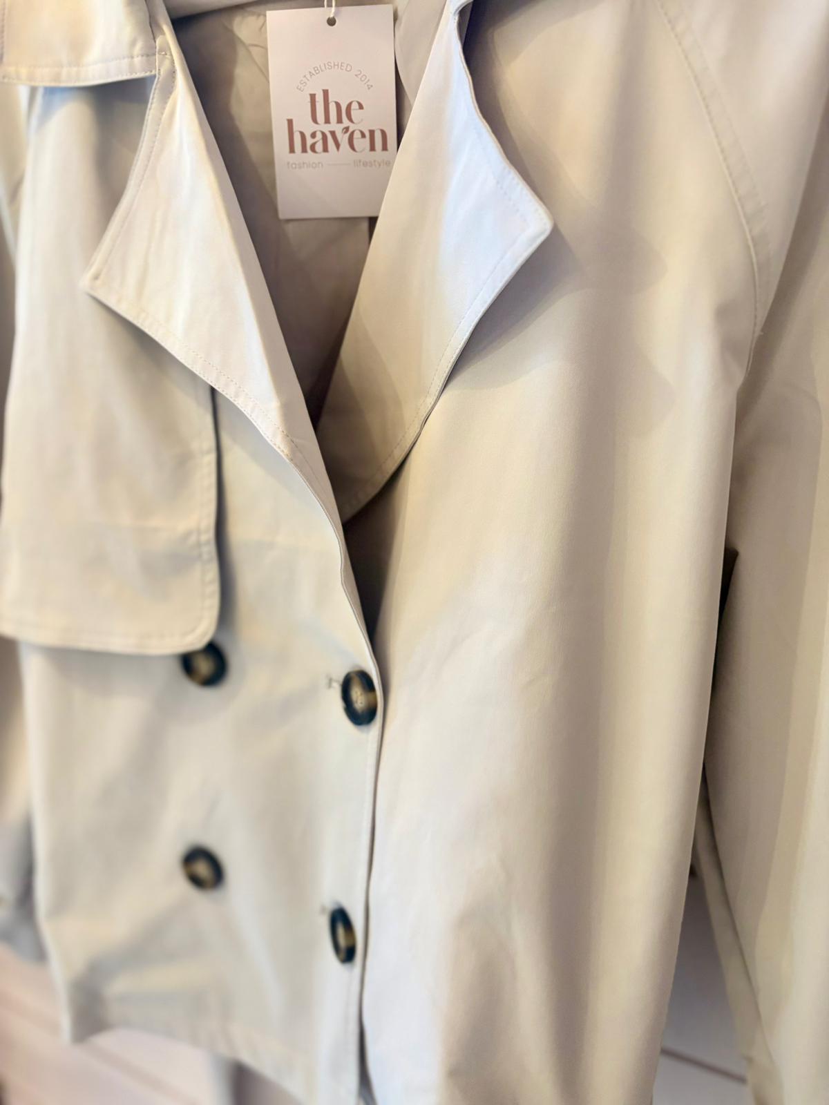 Light-coloured trench coat with black buttons hanging on a rail, featuring a tag labelled "the haven".