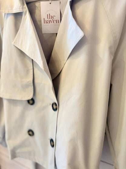 Light-coloured trench coat with black buttons hanging on a rail, featuring a tag labelled "the haven".