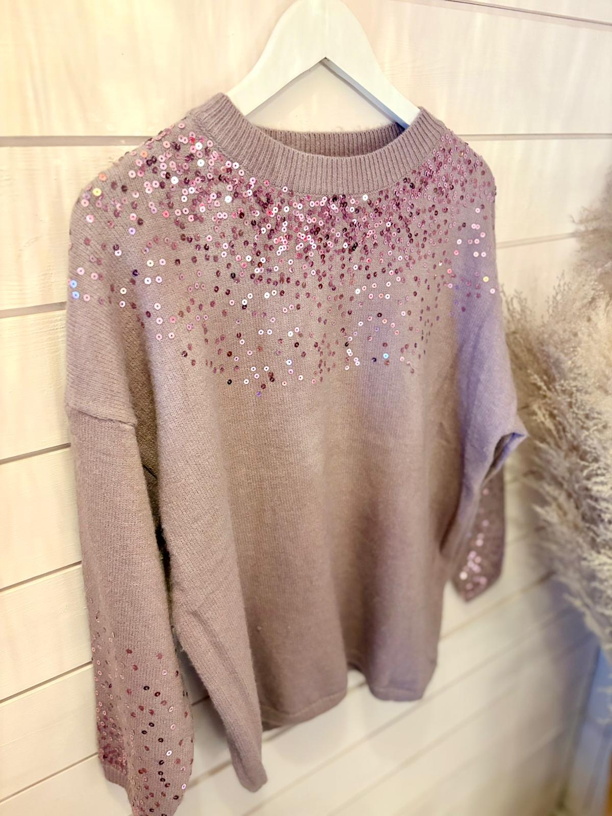 A purple sequin jumper made from vegan materials hangs on a white hanger against a light wooden panel wall, showcasing mauve knit adorned with pink and silver sequins.