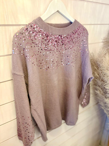 A purple sequin jumper made from vegan materials hangs on a white hanger against a light wooden panel wall, showcasing mauve knit adorned with pink and silver sequins.