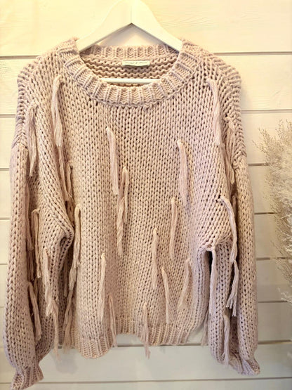 A chunky, light beige tassel jumper with loose, hanging yarn threads is shown on a white hanger against a white wooden wall.