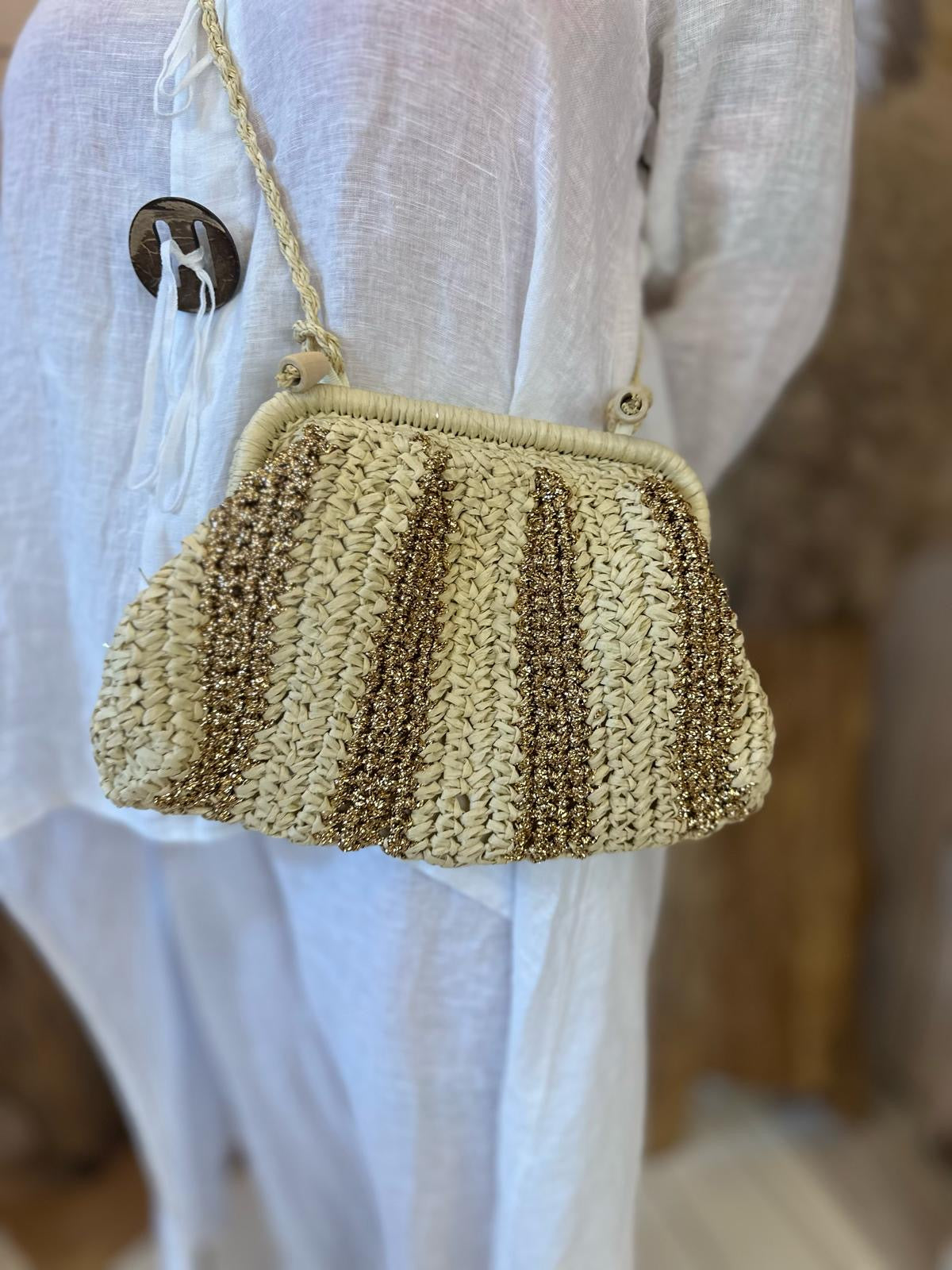 A person wearing a white outfit with large brown buttons is holding an environmentally friendly straw bag with gold accents and a braided strap.