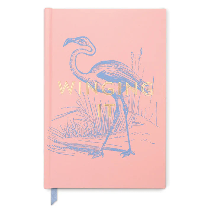 A pink, high-quality notebook with a soft-touch cover, displaying a flamingo illustration and the words WINGING IT in gold lettering on the front.