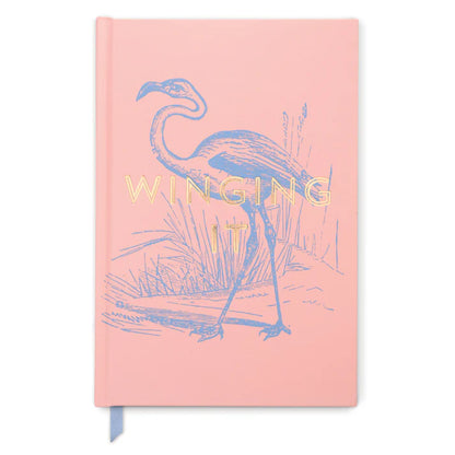 A pink, high-quality notebook with a soft-touch cover, displaying a flamingo illustration and the words WINGING IT in gold lettering on the front.