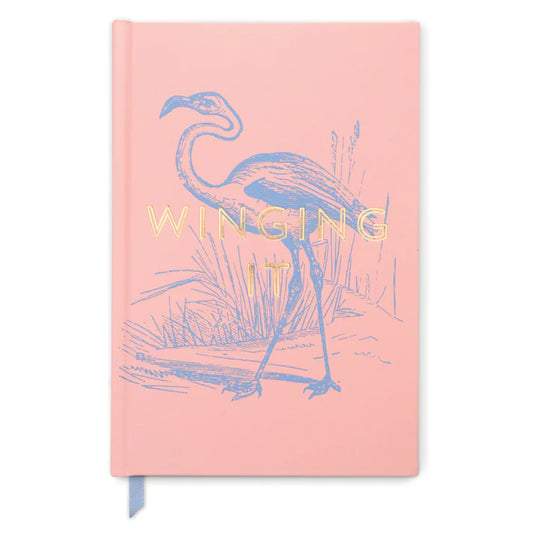 A pink, high-quality notebook with a soft-touch cover, displaying a flamingo illustration and the words WINGING IT in gold lettering on the front.