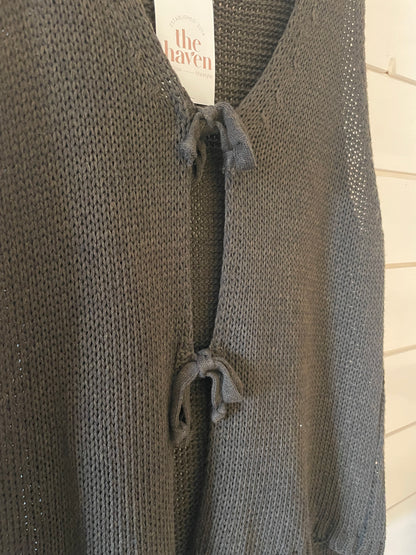 A close-up of a dark grey knitted jumper with two front tie fastenings, ideal for versatile layering, and a The Haven brand label visible at the collar.