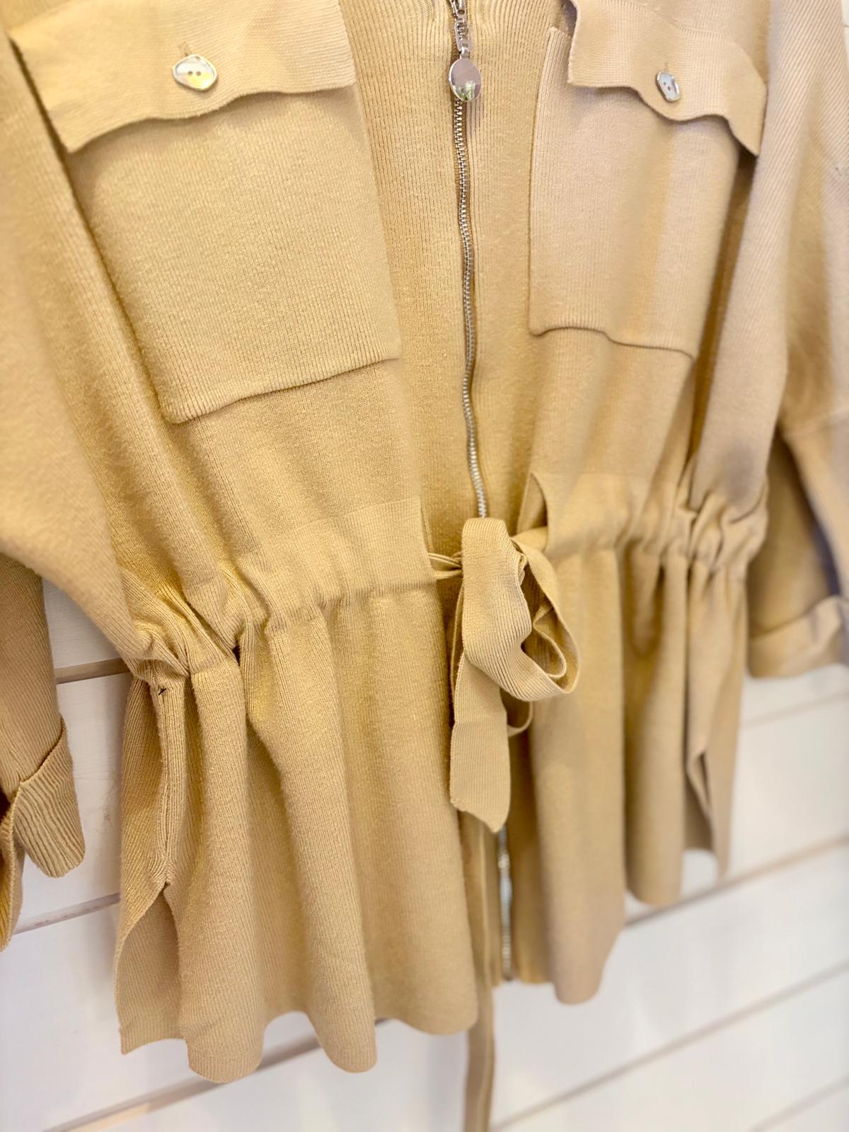 Beige jacket featuring two chest pockets, a front zip fastening, and a fabric belt at the waist, displayed against a white slatted wall.