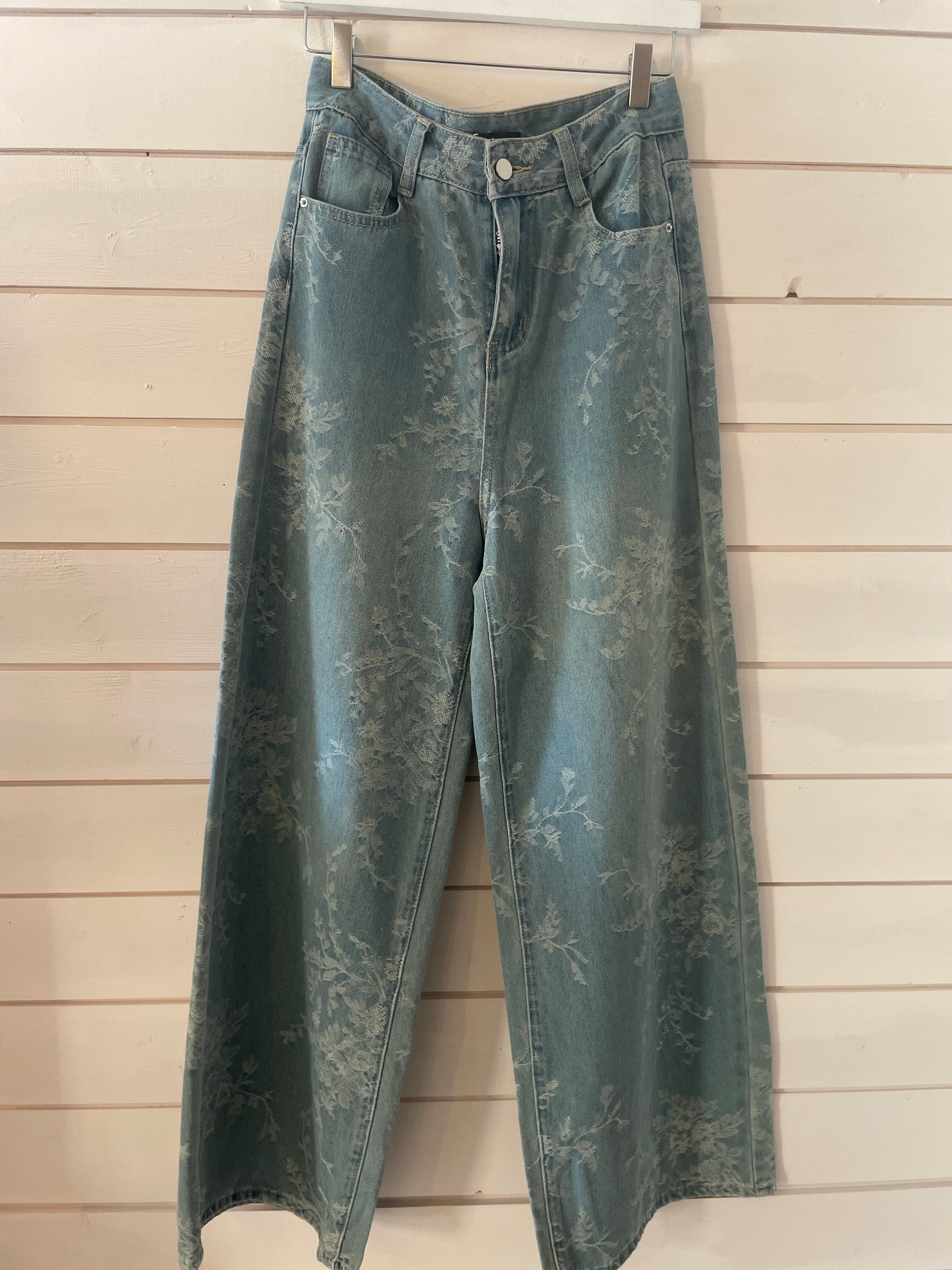 Ultra-soft, wide-leg blue floral jeans with a subtle flower pattern hang on a wooden hanger against a pale wood-panelled wall.
