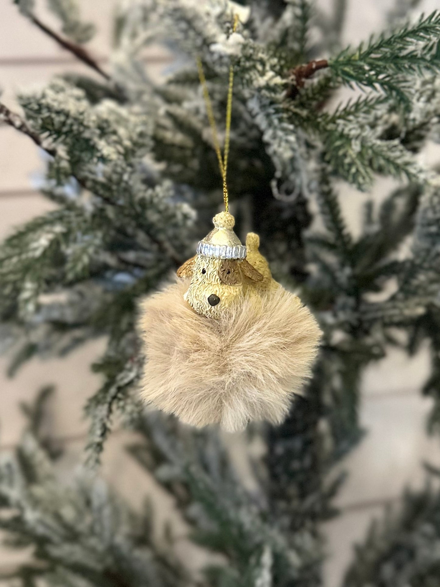 A dog-themed Christmas tree decoration shaped like a fluffy brown sphere with a small dog's head wearing a winter hat, ideal festive décor for dog lovers, hanging from a snow-covered tree branch.