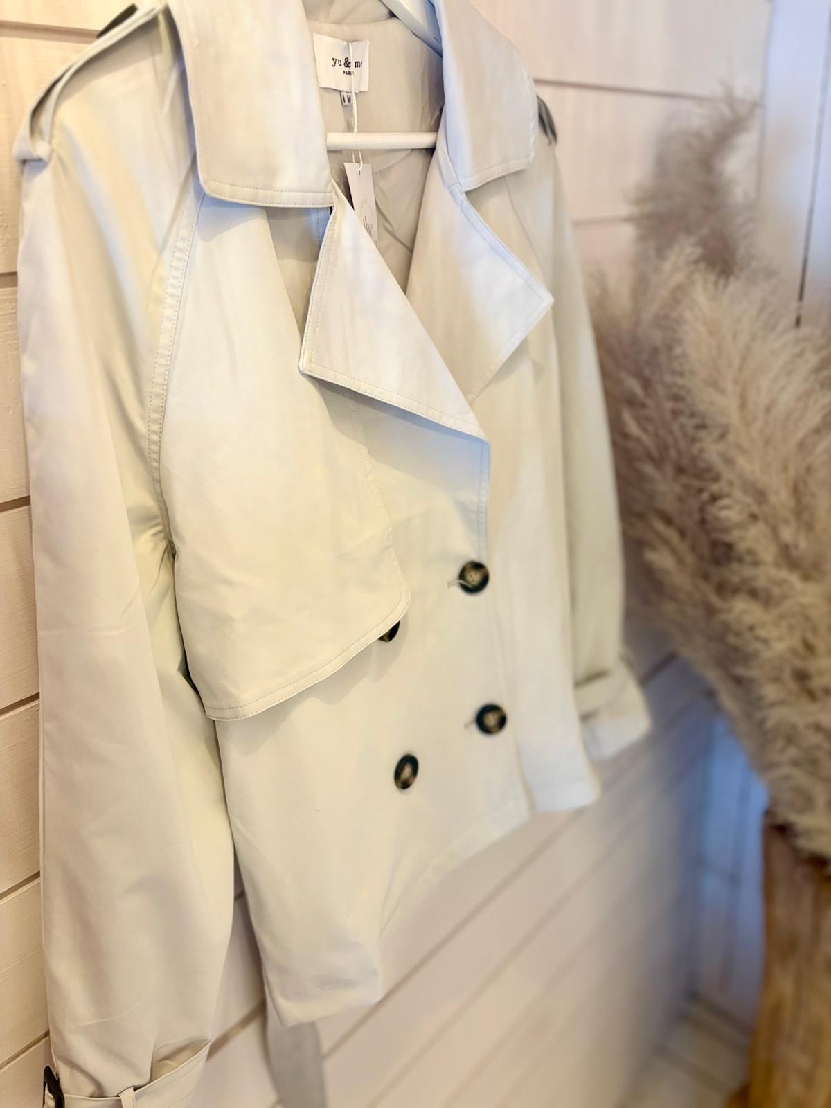 A light beige double-breasted trench coat with black buttons is hanging from a wall-mounted hook beside pampas grass.