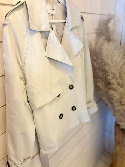 A light beige double-breasted trench coat with black buttons is hanging from a wall-mounted hook beside pampas grass.