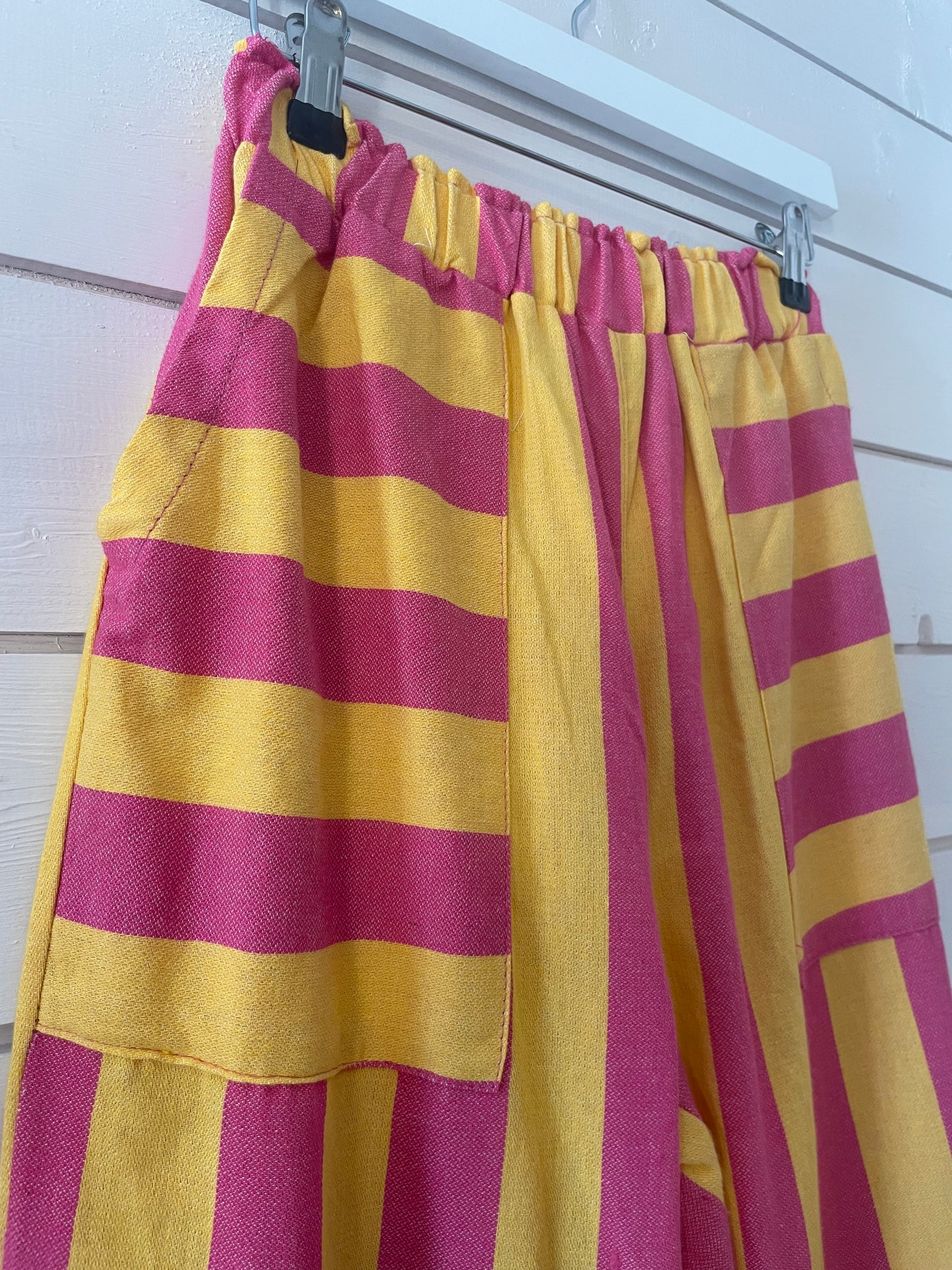 Yellow and pink striped trousers with large front pockets, made from lightweight fabric and featuring elasticated waistbands, hang on a white hanger against a white wooden wall.