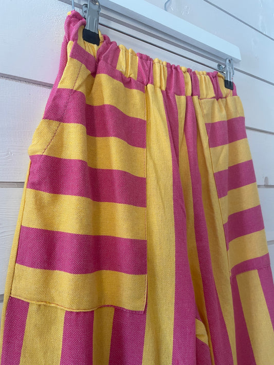 Yellow and pink striped trousers with large front pockets, made from lightweight fabric and featuring elasticated waistbands, hang on a white hanger against a white wooden wall.