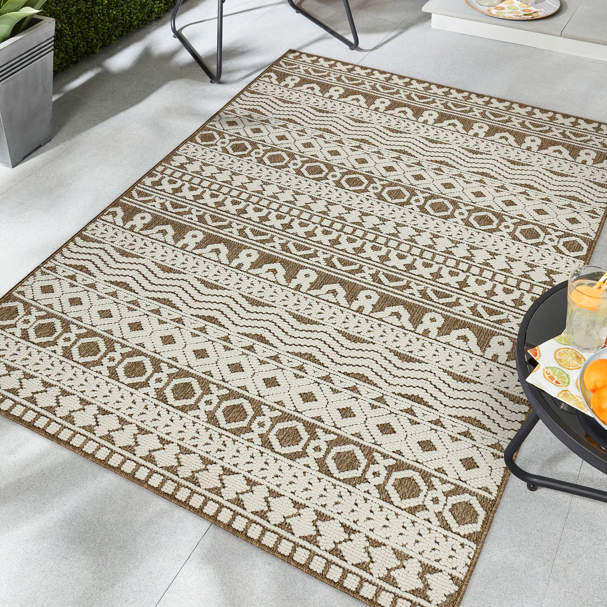 A beige and brown hard-wearing polyester rug featuring geometric and animal designs lies on a light tiled patio beside a small table set with drinks and snacks.