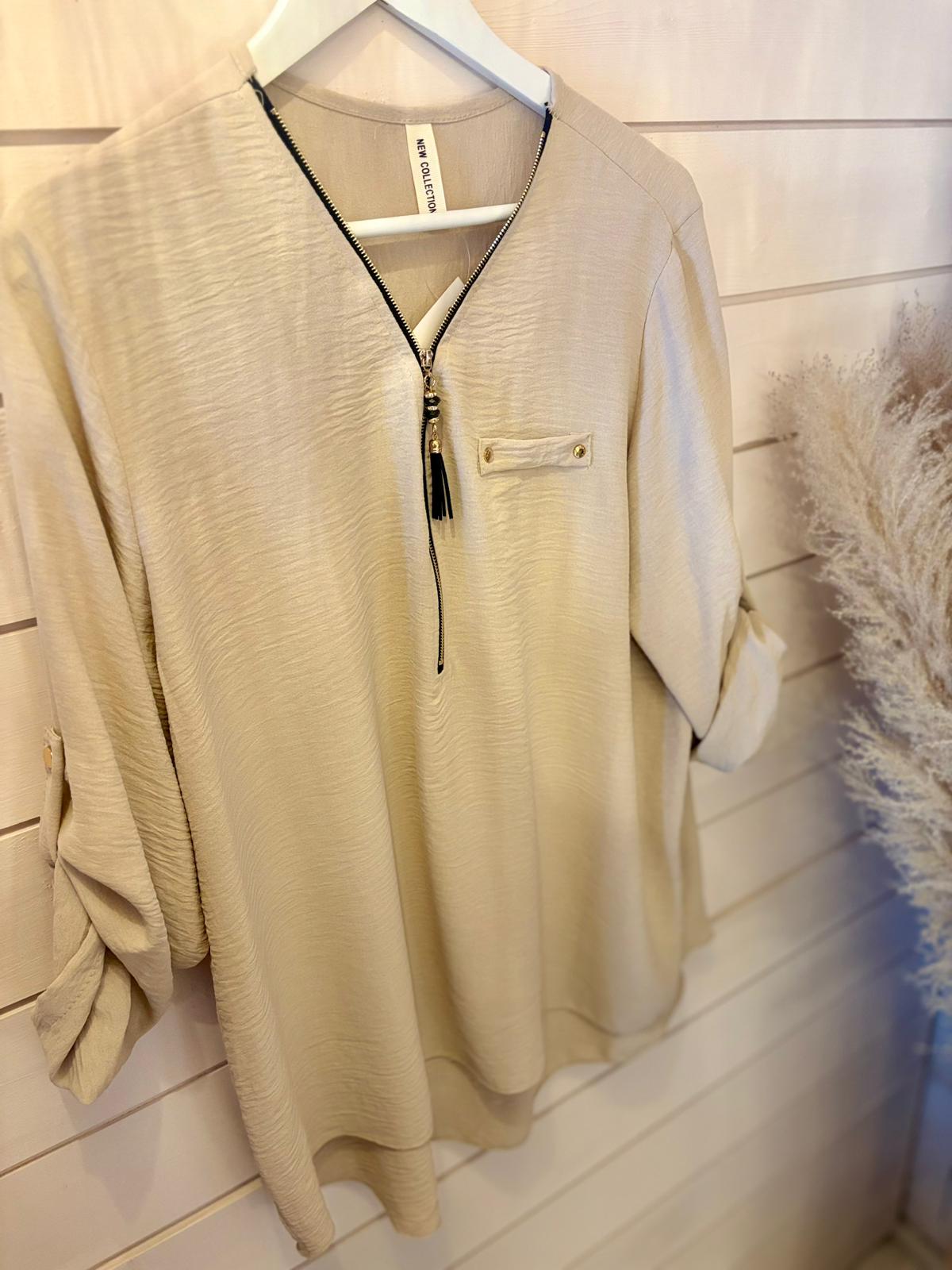 Beige long-sleeved blouse featuring a front zip detail and chest pocket, shown on a hanger against a slatted wall with decorative dried pampas grass nearby.