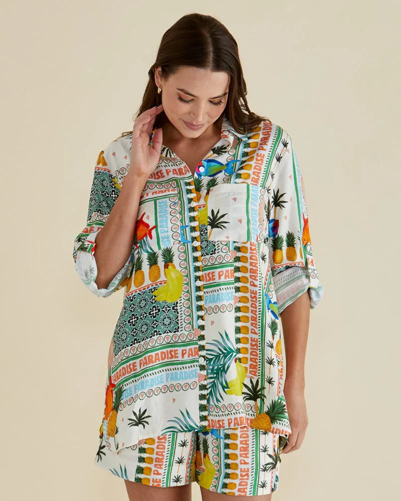 Woman dressed in a matching Paradise Print shirt and shorts set featuring colourful tropical fruits, made from vegan materials, standing against a plain background.