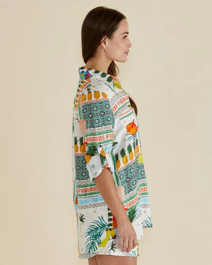 Woman in profile wearing a colourful Paradise Print shirt featuring tropical and geometric patterns, made from vegan materials, standing against a plain light background.