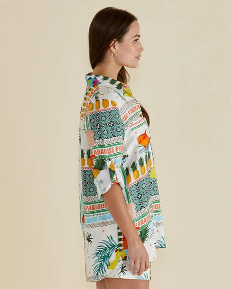Woman in profile wearing a colourful Paradise Print shirt featuring tropical and geometric patterns, made from vegan materials, standing against a plain light background.