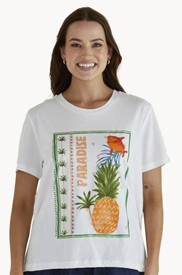 A woman wearing a white T-shirt with a graphic of a pineapple, fish, green plants and the word PARADISE printed vertically, made from breathable fabric for relaxed coastal style.