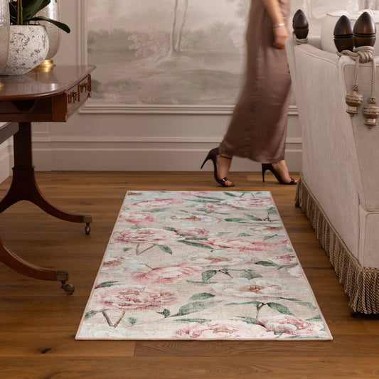 A woman in a long dress walks past a rug patterned with blooming peonies in a room with wooden flooring, a sofa, and a table with a plant.