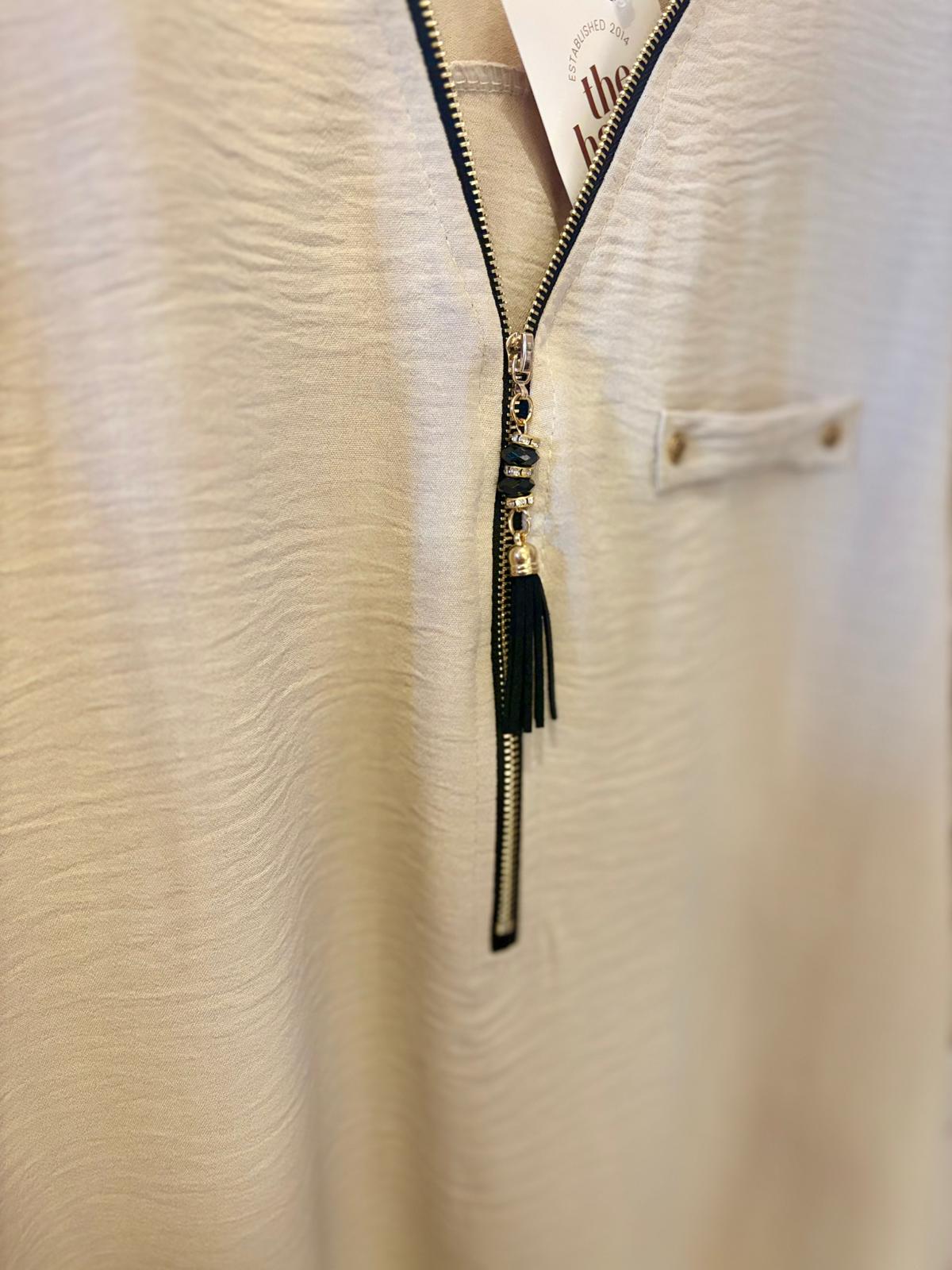 Close-up of a beige textured shirt featuring a half-zip neckline, black zip pull with a tassel, and a small chest pocket with a button.