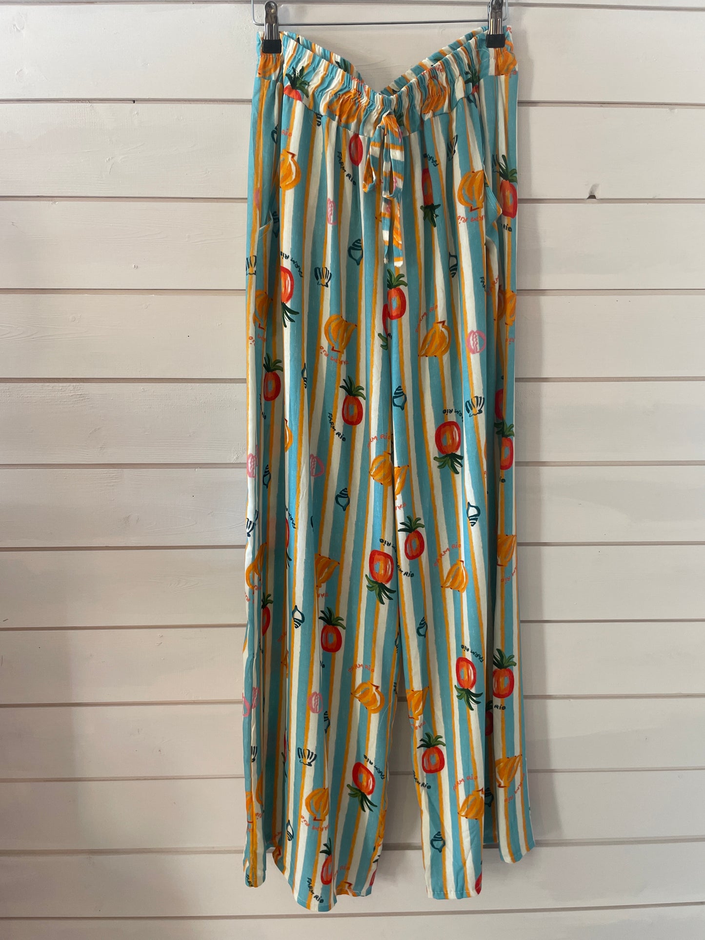 Wide-leg, lightweight trousers with blue and white stripes and playful fruit prints, made from vegan-friendly fabric—ideal for casual wear, shown against a white wooden wall.