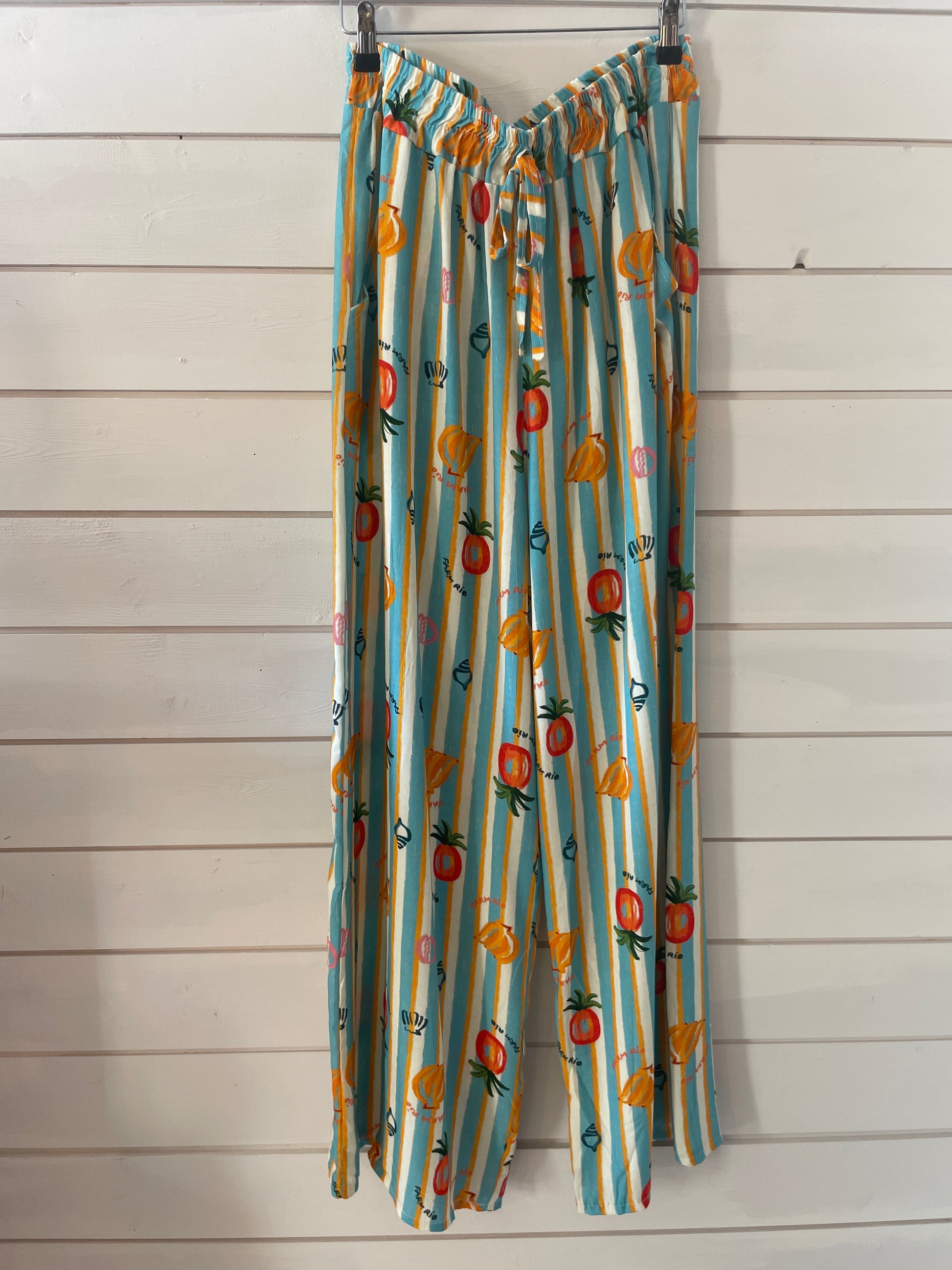 Wide-leg, lightweight trousers with blue and white stripes and playful fruit prints, made from vegan-friendly fabric—ideal for casual wear, shown against a white wooden wall.