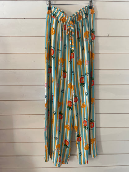 Wide-leg, lightweight trousers with blue and white stripes and playful fruit prints, made from vegan-friendly fabric—ideal for casual wear, shown against a white wooden wall.