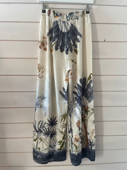 Blue jungle trousers in lightweight fabric with an elasticated waistband, showcasing a botanical print in navy, green, and brown on a cream base, hung on a clear hanger against a white wooden wall.