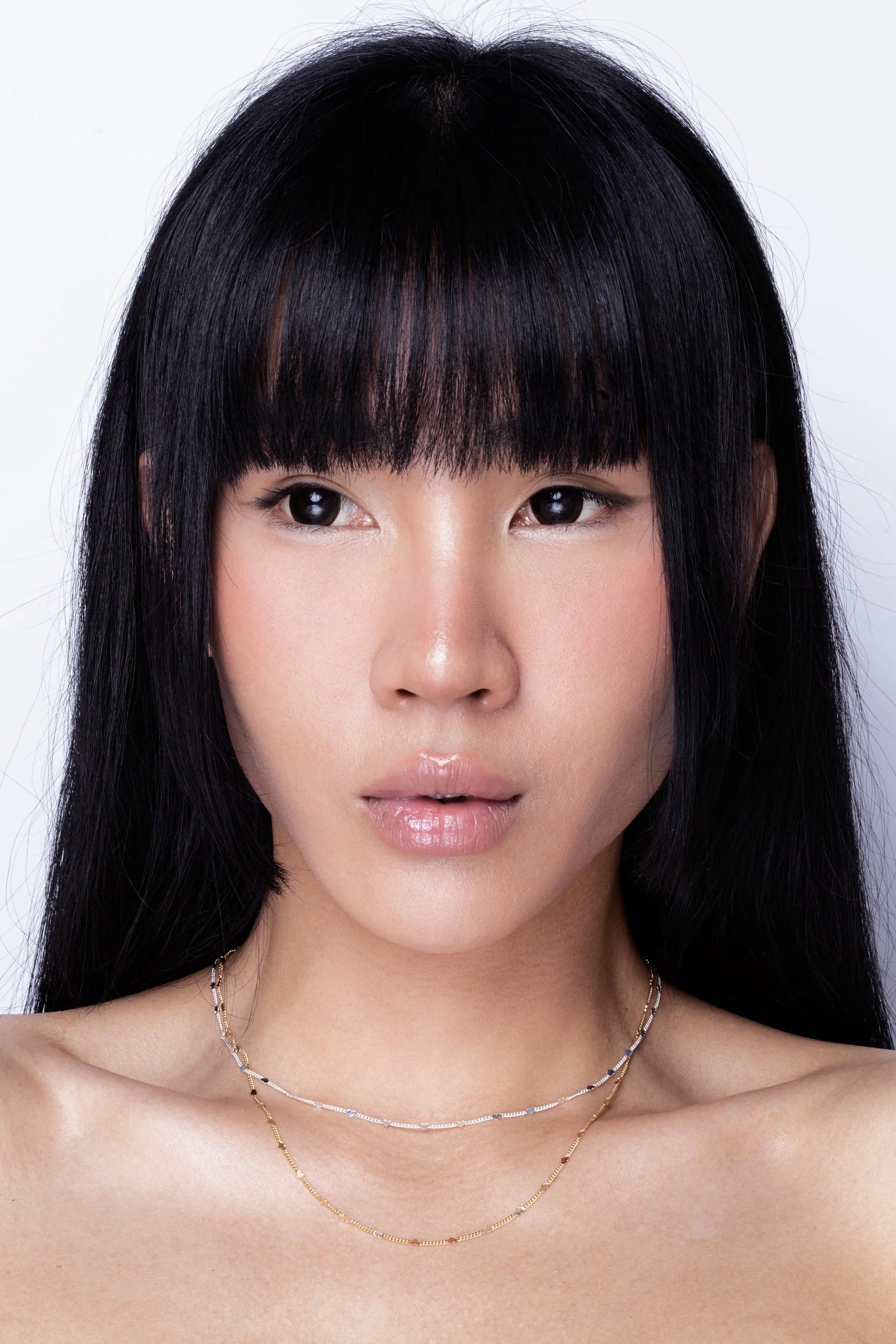 A woman with straight black hair and a fringe wears a delicate hypoallergenic necklace and a gold chain against a plain white background.